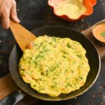 Suji Chilla Recipe: A golden-brown, thin pancake cooking in a black skillet with a wooden spatula. A small bowl of dipping sauce is on the side.