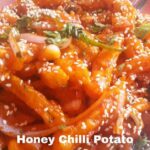 Close-up shot of a plate filled with Honey Chilli Potato. The dish features crispy fried potato strips coated in a glossy orange sauce, garnished with white sesame seeds, chopped onions, and fresh cilantro. The text "Honey Chilli Potato" is overlaid in white at the bottom of the image.