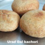 Close-up shot of three golden brown Urad Dal Kachoris on a silver plate. The kachoris are round and slightly flattened, with a textured surface.