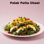 A plate of Palak Patta Chaat on a pink background. The dish features crispy spinach leaves topped with yogurt, sev (thin fried noodles), pomegranate seeds, and other garnishes.