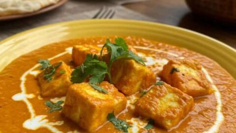 Recipe of Shahi Paneer: A yellow plate with shahi paneer, featuring paneer cubes in a creamy orange sauce, garnished with cilantro and a swirl of cream. Other dishes and naan bread are visible in the background.