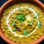 Green Moong Dal Recipe: A close-up, overhead shot of a bowl of green moong dal (mung bean curry) in a terracotta serving dish. The dal is a vibrant yellow-green color with visible whole green mung beans. It is topped with a swirl of white cream, a pat of melting butter, fresh coriander leaves, and a small dried red chili.
