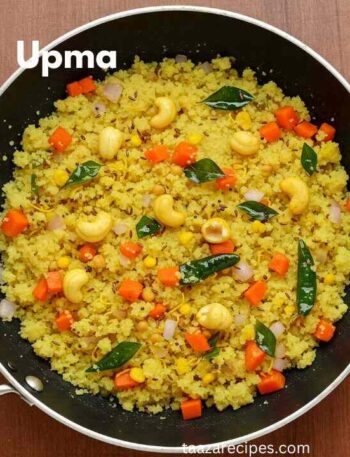 Recipe For Making Upma: A top-down view of a pan filled with cooked upma, a South Indian savory semolina dish. The upma is yellow, speckled with diced carrots, white onions, cashew nuts, and curry leaves. The pan is black with a silver handle, and the dish is presented on a wooden surface.